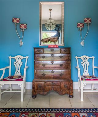 Blue entryway with antique chest of drawers, white armchairs and tiled floor