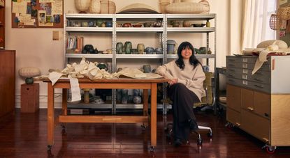 A young woman dressed in an oversized beige jumper and dark trousers smiles while sitting at a big wooden working desk surrounded by paper lanterns and ceramic creations.