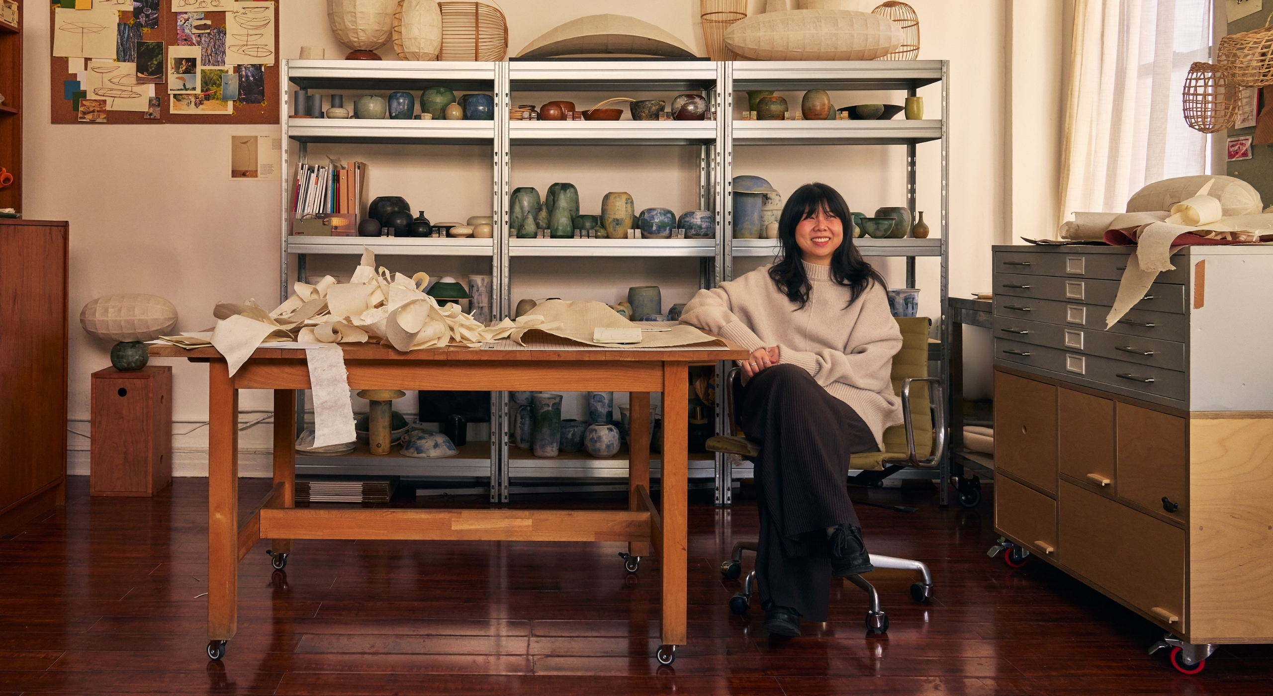 A young woman dressed in an oversized beige jumper and dark trousers smiles while sitting at a big wooden working desk surrounded by paper lanterns and ceramic creations.