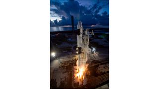 a black and white spacex rocket test-fires its first-stage engines on the launch pad beneath a dark-blue late-evening or early-morning sky, with a body of water in the background