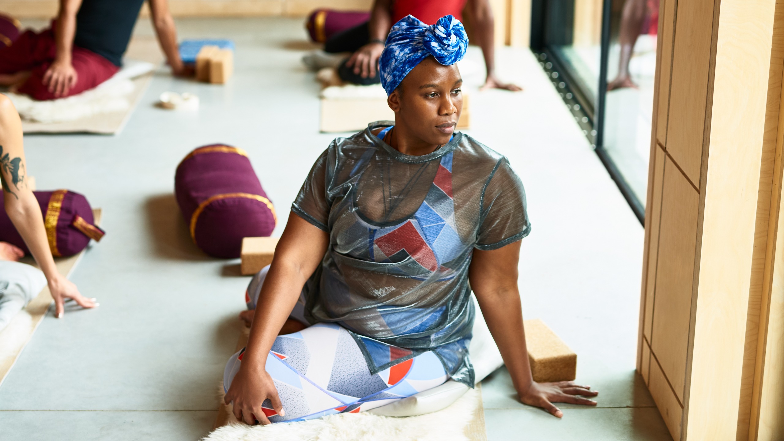 A woman in a yoga class sits on the floor with her legs crossed, her right hand on her left knee, and her left hand behind her body. She is twisting her torso to the left.