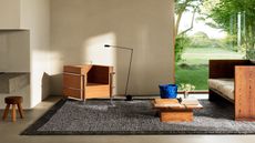 Image of a modern living room with white walls and gray concrete flooring. There is a gray and black short pile shag rug on the floor and a set of matching wooden seating (a sofa and accent chair). There is also a low to the ground, wooden coffee table with coffee table books on it.