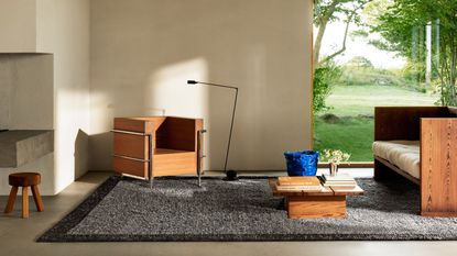 Image of a modern living room with white walls and gray concrete flooring. There is a gray and black short pile shag rug on the floor and a set of matching wooden seating (a sofa and accent chair). There is also a low to the ground, wooden coffee table with coffee table books on it.