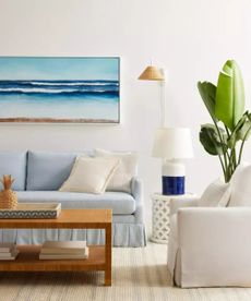 A pale blue sofa with white throw pillows and a white armchair around a wooden coffee table; in the background, white walls and a landscape painting of the sea. 