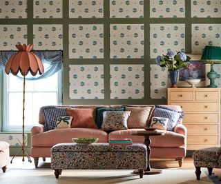 square wooden wall panels on wall with floral blue and cream wallpaper inset in panels, pink sofa, pink floor lamp, floral patterned ottoman and light wooden chest of drawers