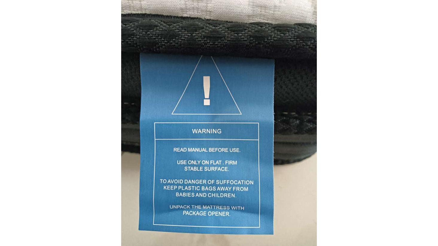 A blue warning label on the side of the Crayan Mattress. The label advises to read the manual before use, use on a flat stable surface, warns about suffocation risk and advises how to unpack the mattress