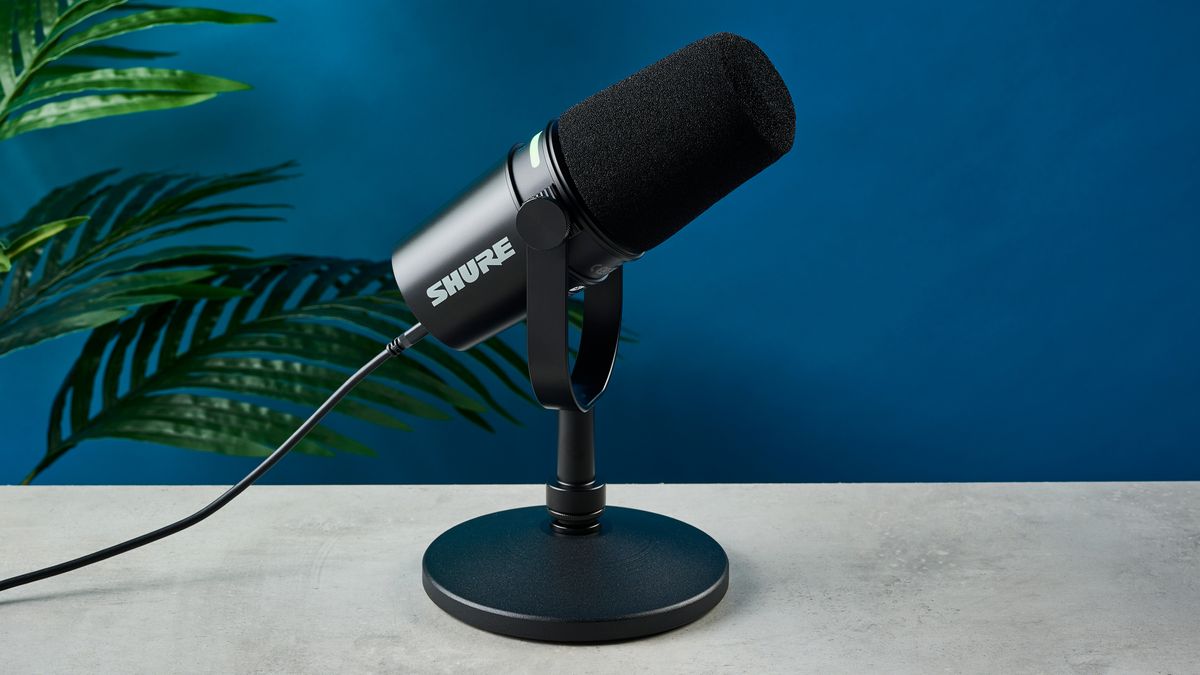 a black microphone with a desktop stand and a black muff is on top of a beige surface with a blue backdrop