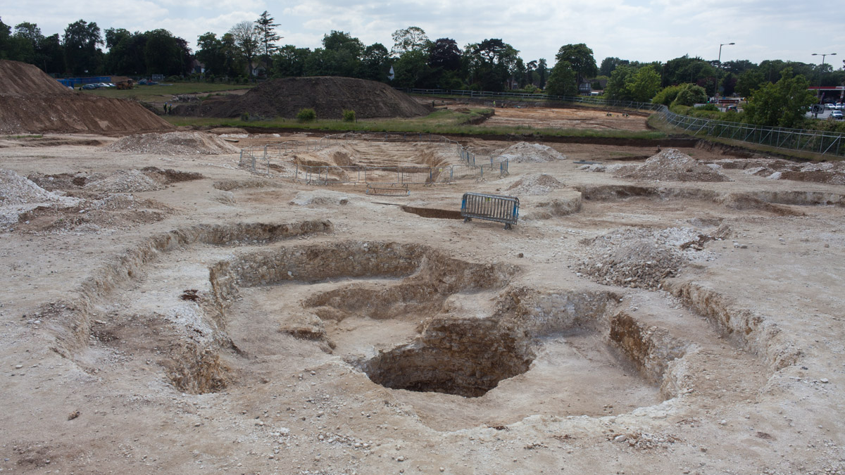 View of a shaft being excavated in Surrey, England. The foreground is light brown with concentric circles stepped into the dirt. In the background is a ring of trees against a cloudy blue sky.