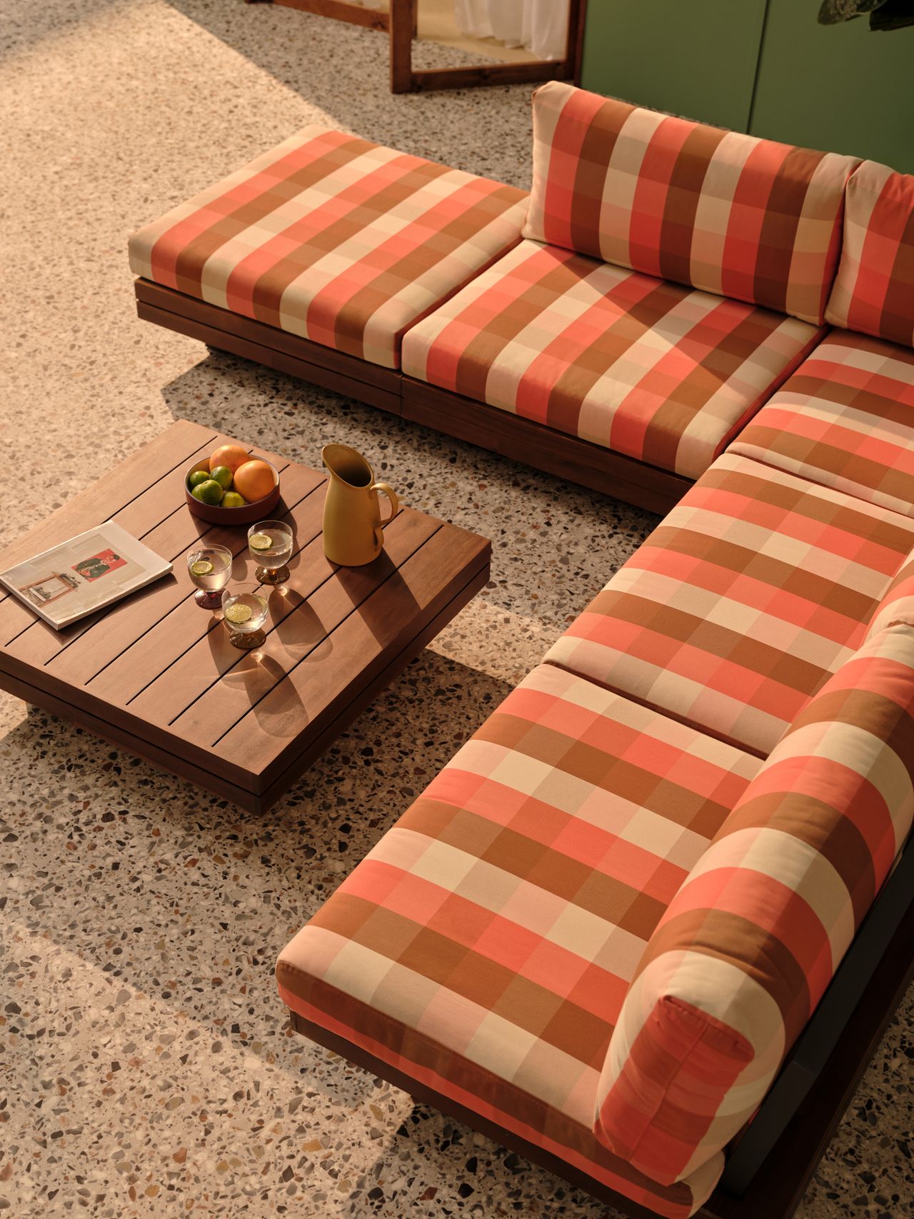 A pink, brown, and white check outdoor sectional sofa with a brown wooden coffee table in front of it. THe floor is made of terrazzo tile.
