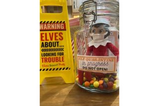 Elf on the Shelf wearing a face mask and pictured sitting intside a jar surrounded by sweets and holding a note explaining that he is in quarantine due to Covid travel restrictions
