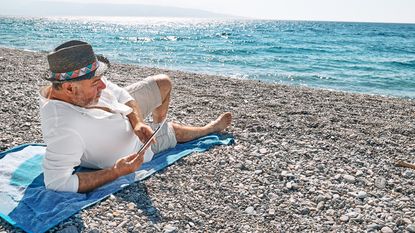 An older man relaxes on the beach with a tablet.