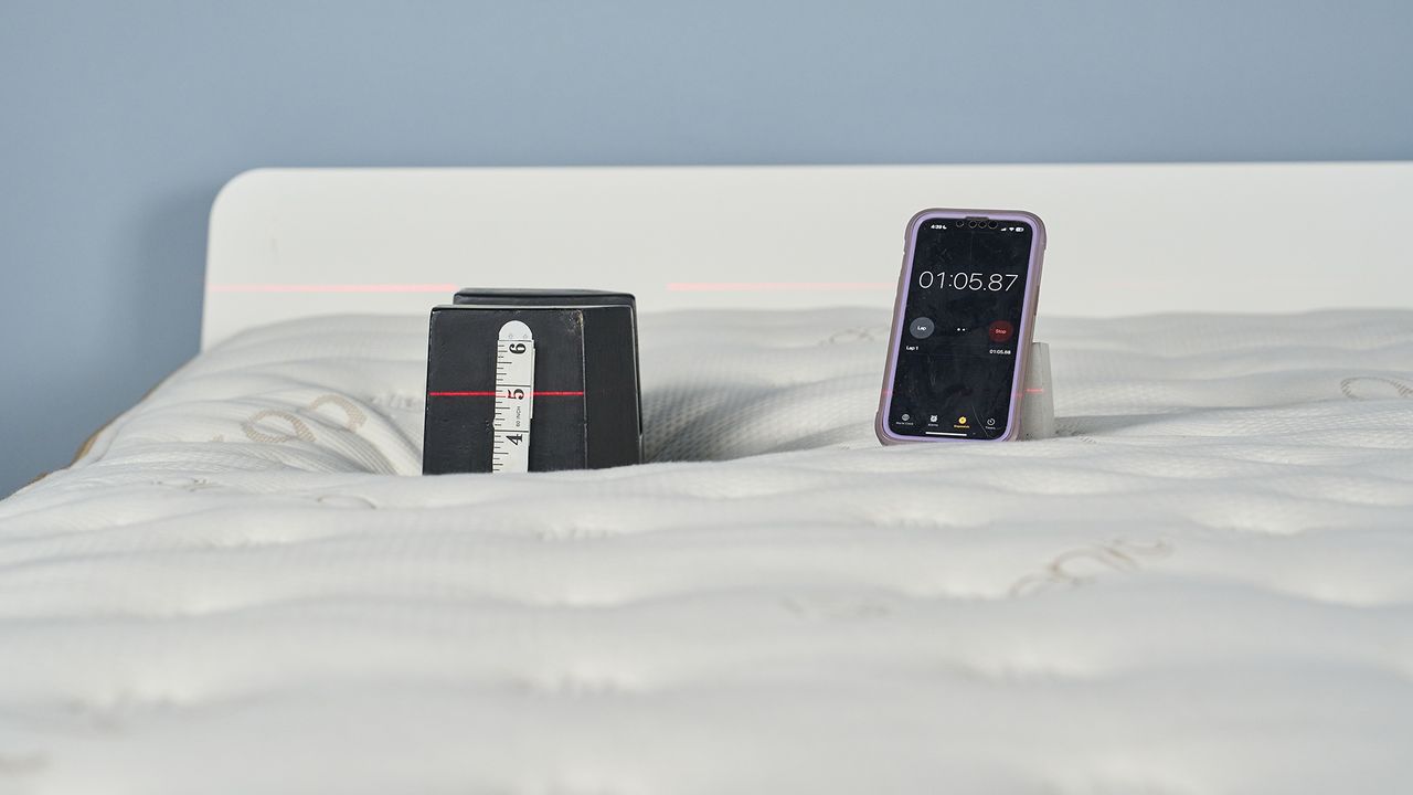 Image shows the highest weight in our pressure relief weighted indentation test being placed on top of the Saatva Classic: a 25kg cast iron weight placed in the shoulder area, which is a key impact zone on the body when side sleeping and back sleeping. A measuring tapes gives us a visual record of how much the mattress is indenting at that spot under the 25kg weight.
