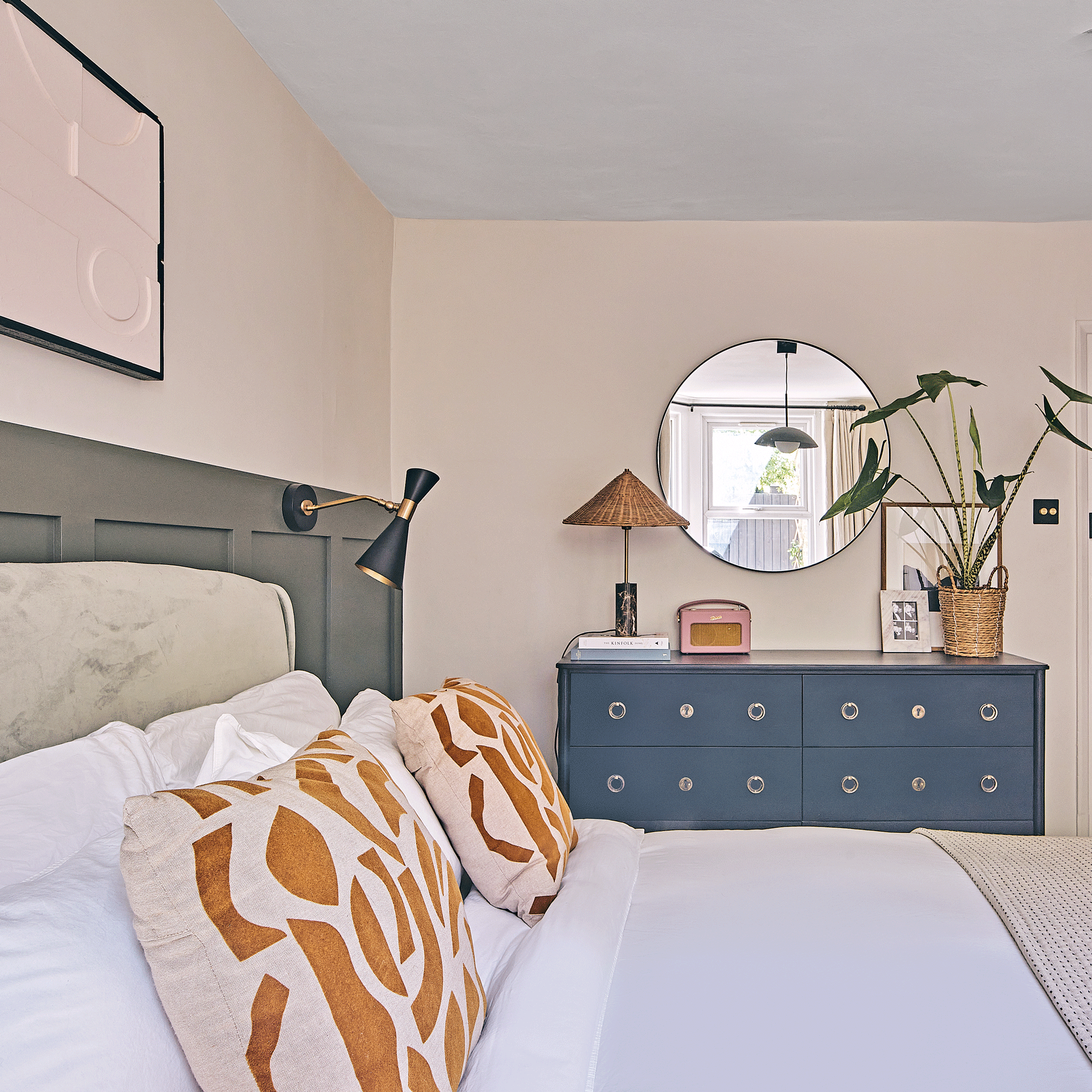 a bedroom with dark green wall panelling a dark blue chest of drawers and a round wall-hung mirror