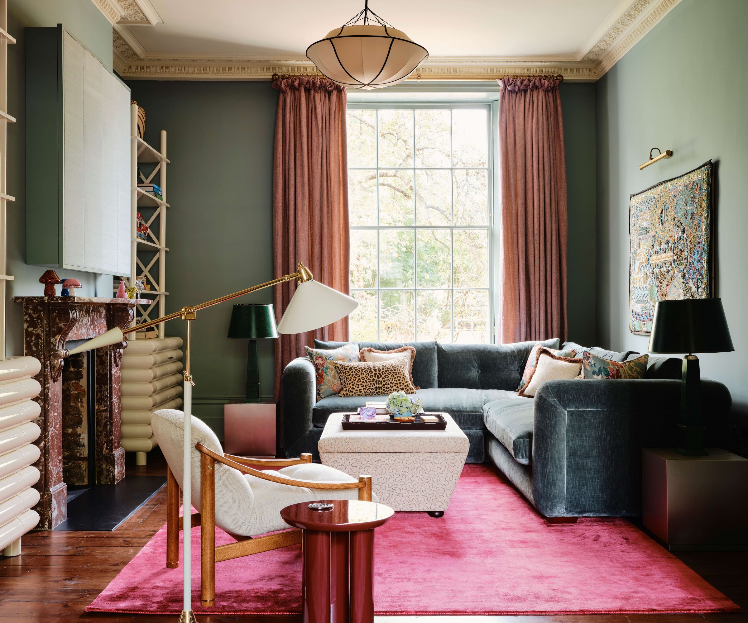 blue living room with pink curtains and a pink rug with a deep velvet blue corner sofa, ottoman, wooden armchair, and side table with lamp