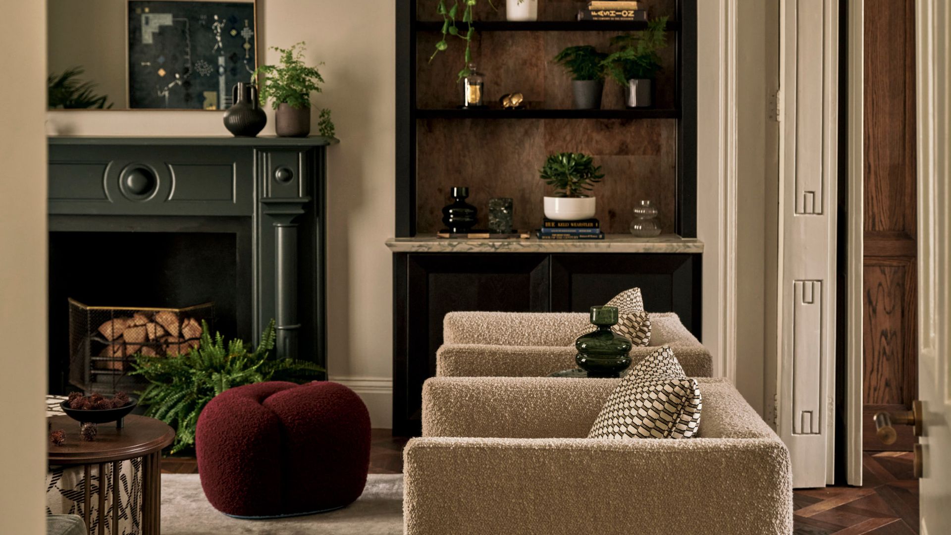 A beautiful living room with a green mantel over a fireplace, a maroon pouf, a wood coffee table, a pair of cream boucle armchairs, patterned cushions, and potted houseplants like jade, fern, and trailing plants