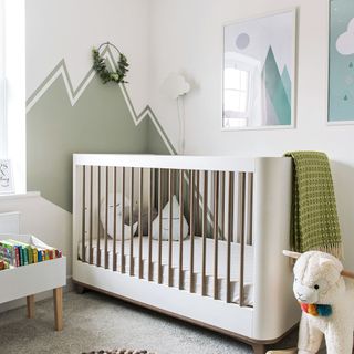 White nursery with green painted mountain mural behind a white cot