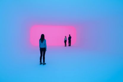 James Turrell, Aftershock Light & Space features three people (one standing back) looking towards a purple light. 