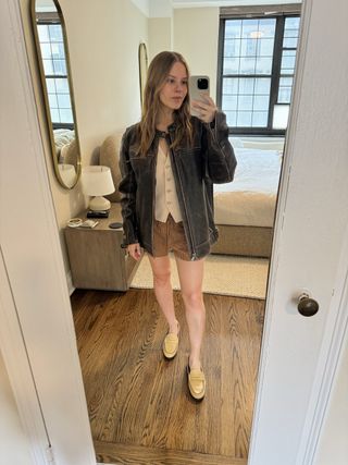 Nikki Chwatt wears a brown leather jacket, a beige tailored vest, brown striped shorts, and beige Koio loafers.