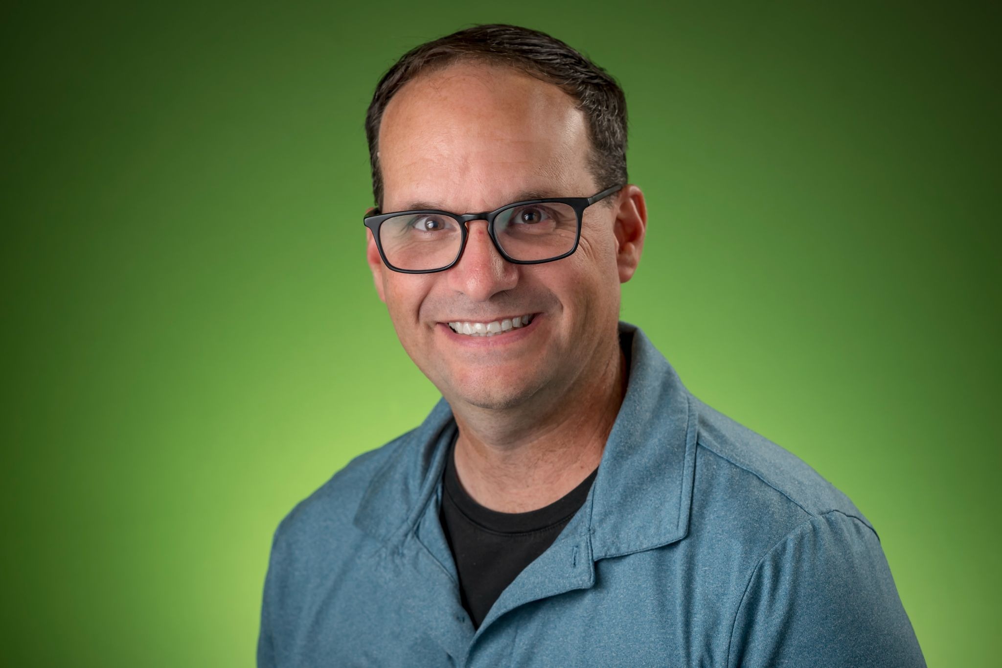 man smiling at the camera against a green background wearing a blue shirt and black glasses.