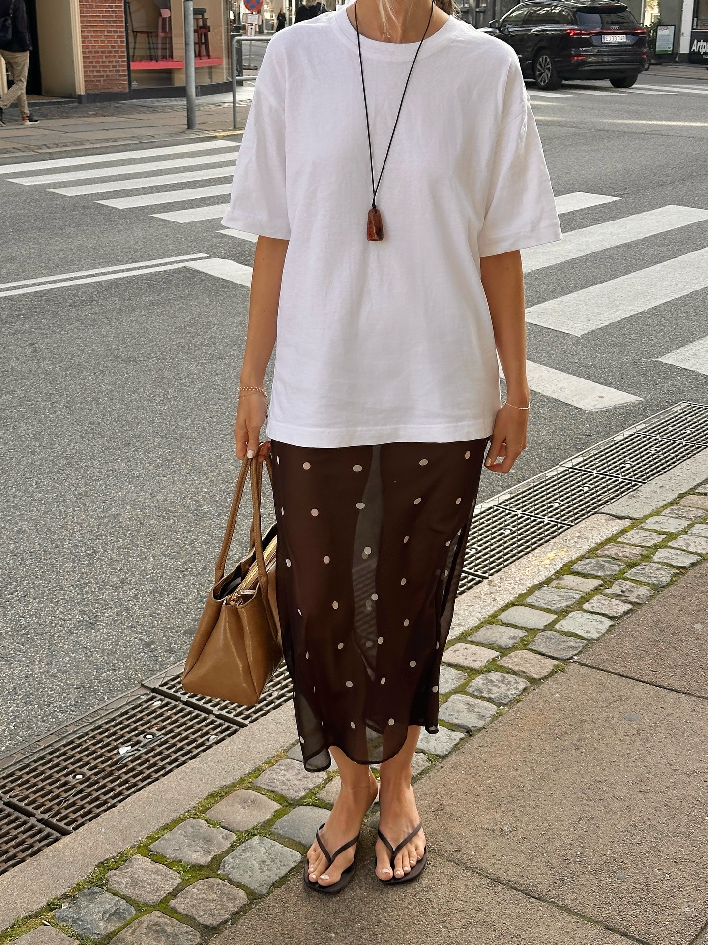 British influencer Chloe Kath Butler poses on a London sidewalk wearing an oversize white T-shirt, a long pendant necklace, a camel bag, brown sheer polka-dot print skirt, and brown flip-flop sandals