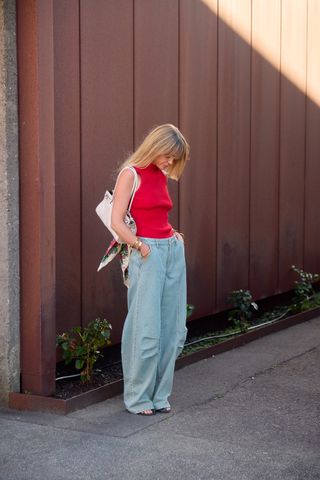 A woman wearing a red tank top with light wash baggy jeans, sandals, and a white shoulder bag that has a silk scarf tied to it.