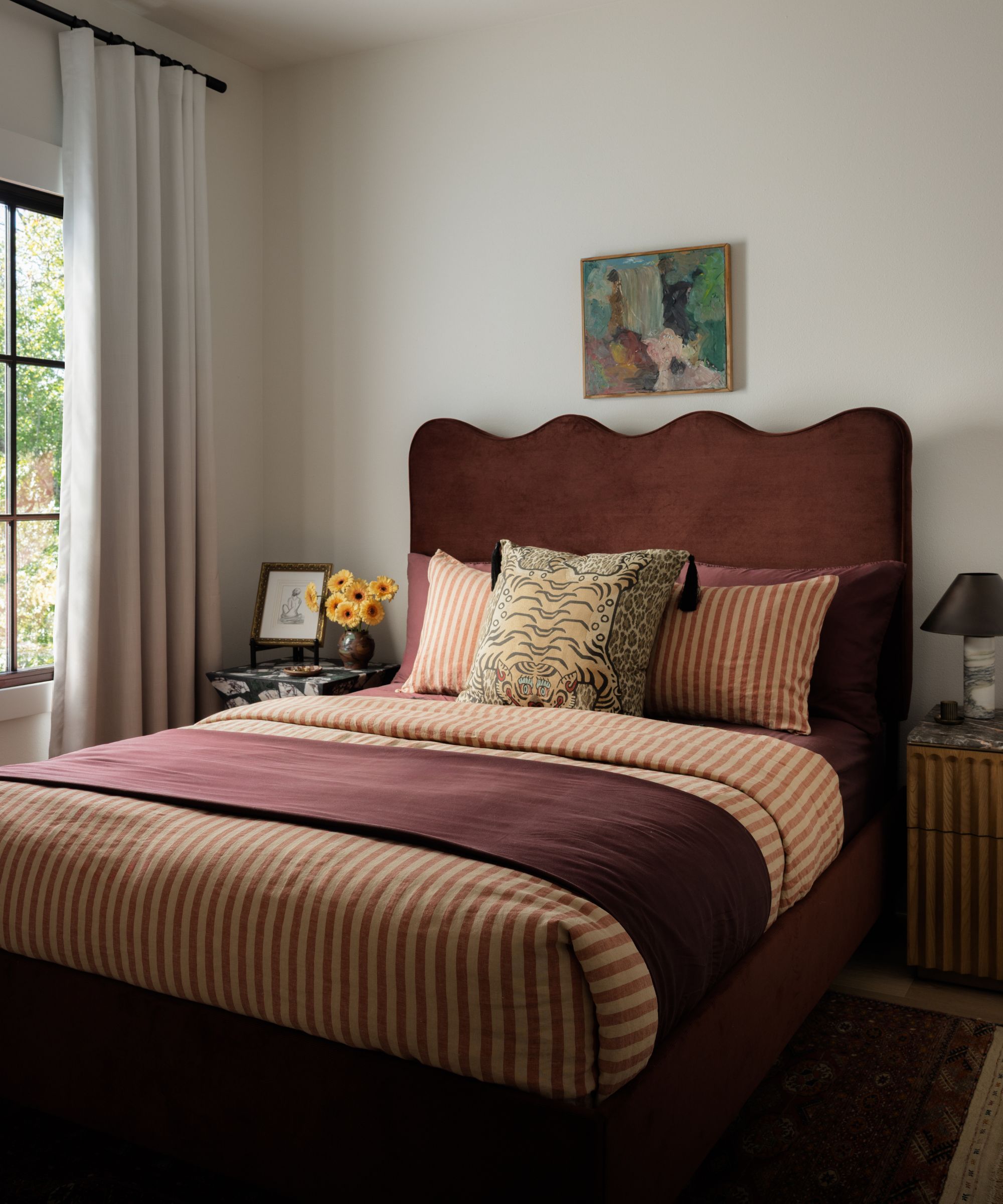 a neutral bedroom with large window and a maroon velvet bed with wavy shape, striped bed linen and a house of hackney tiger pillow