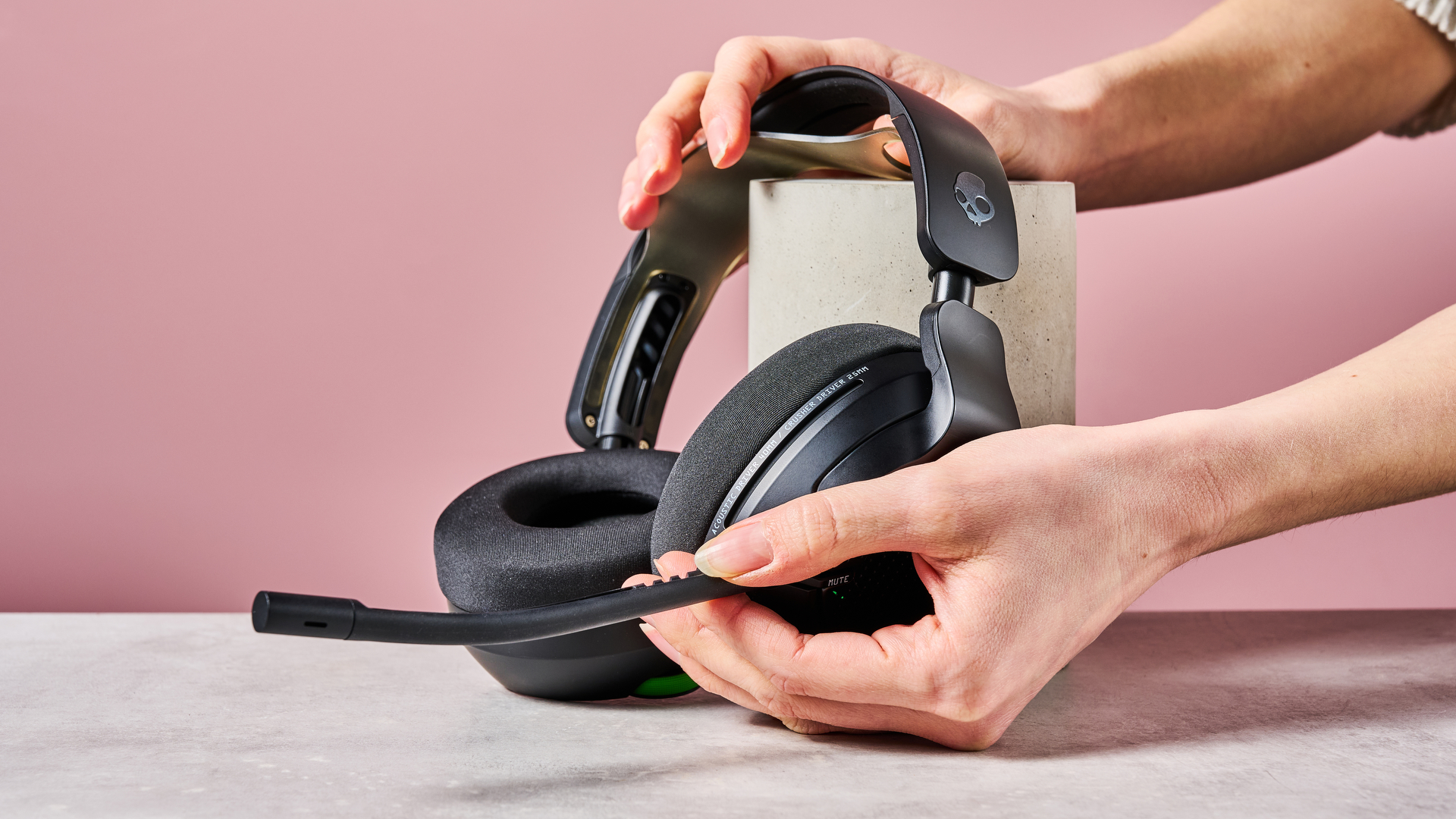 Hands holding Skullcandy Crusher PLYR 720 headset and attached microphone, leaning on a plinth on desk against a pink background