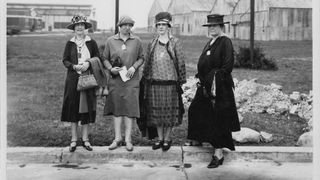 Four women stand on a curb in a black and white photo.