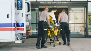 an ambulance parked outside an emergency department. Two paramedics are wheeling a patient on a stretcher away from the ambulance and towards the hospital