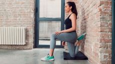 Woman half kneels facing away from an interior exposed-brick wall. Her rear foot is sandwiched between her glute and the wall.