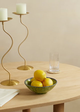 A wooden dining table with a napkin, two curvy gold candle holders, a glass on a coaster and a serpentine bowl full of lemons