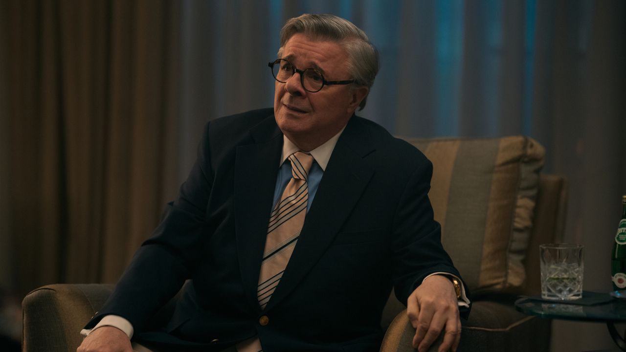 Nathan Lane as Dominick Dunne, sitting in an armchair with a glass on a table next to him, in episode 207 of Monsters: The Lyle And Erik Menendez Story.