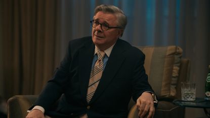 Nathan Lane as Dominick Dunne, sitting in an armchair with a glass on a table next to him, in episode 207 of Monsters: The Lyle And Erik Menendez Story.