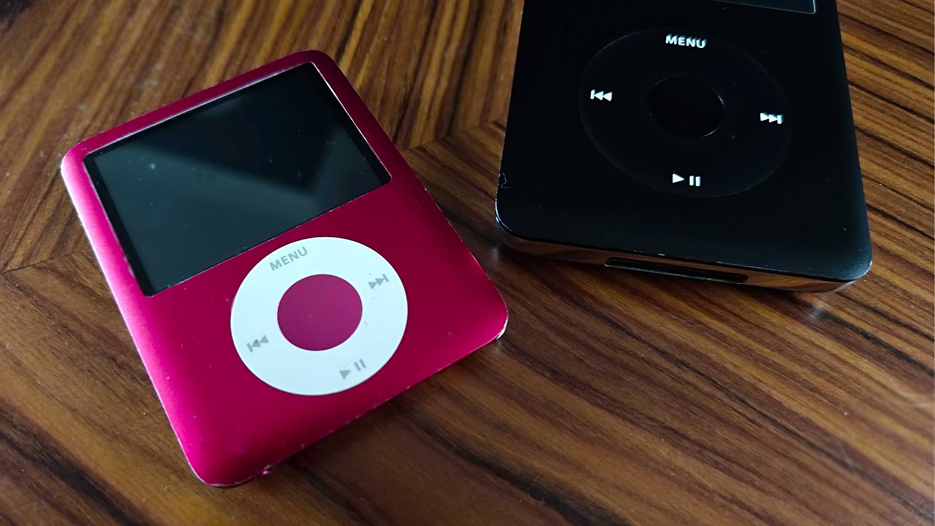 Red iPod Nano 3rd gen next to black iPod classic on woodgrain table.