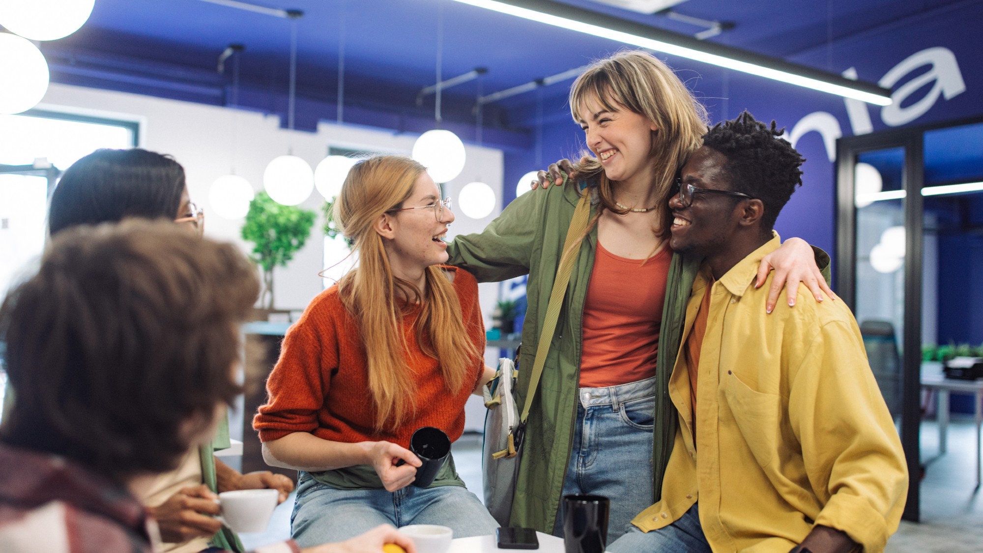 A young woman with her arms around the shoulders of two co-workers in a modern office break room