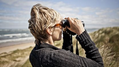 A woman uses binoculars at the beach to look for something. 