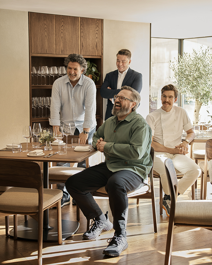 Four middle-aged male restaurateurs, including a chef, sitting on the right-hand side, smile photographed sideways while wearing casual-smart clothing in a linearly designed restaurant with wooden furnishings and bathing in natural light.
