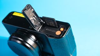 An upside down Fujifilm X100VI mirrorless camera against a blue background with the battery hatch open.
