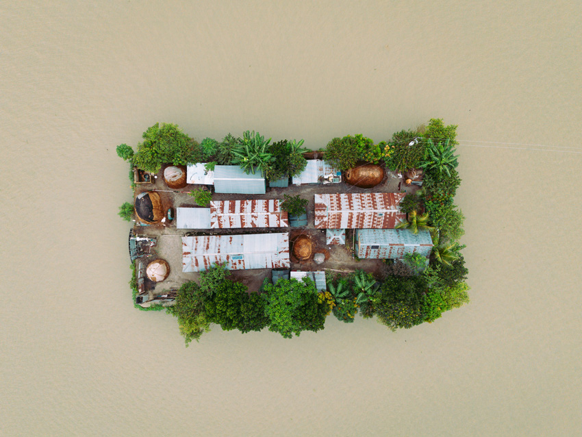 Aerial view of a small, lush green island surrounded by water, featuring tin-roofed buildings and circular thatched structures