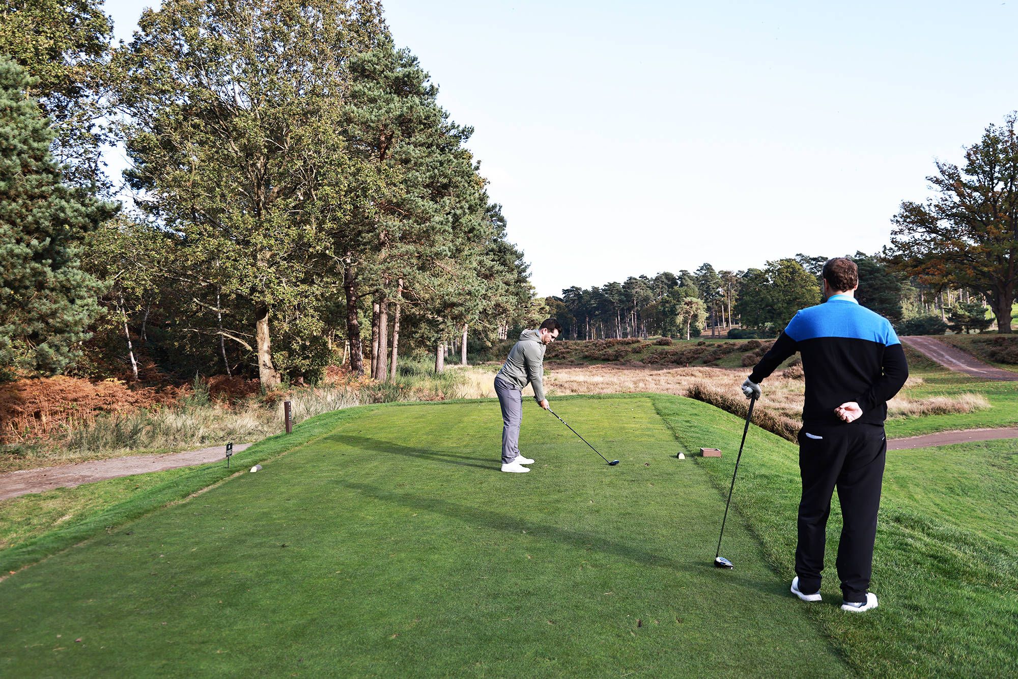 Sam De'Ath hitting 3-wood off the tee, watched on by Nick Bonfield on the tee box holding his driver in anticipation of his next tee shot