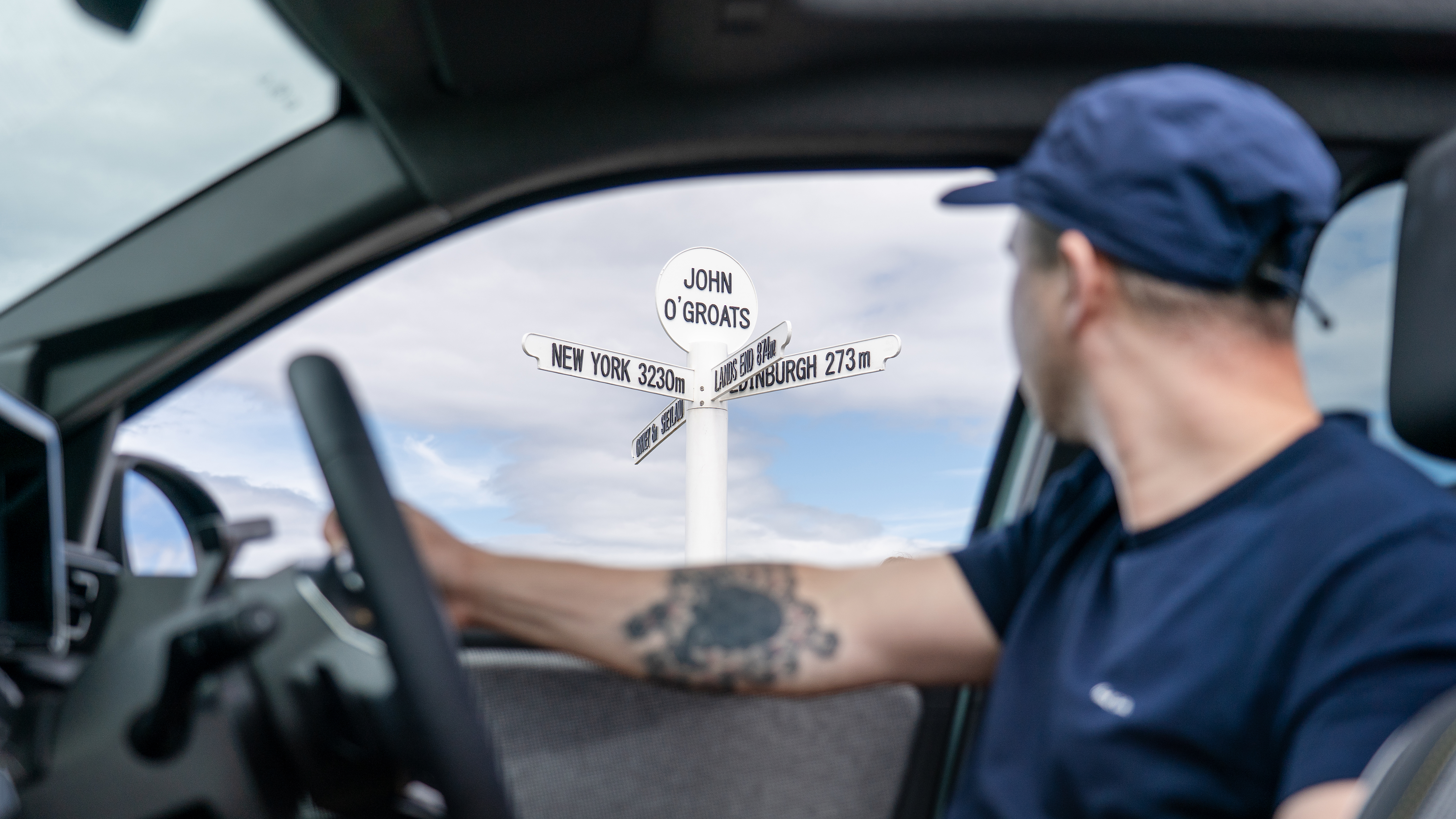 A man looking out of the window of an EV at a signpost for John o'Groats
