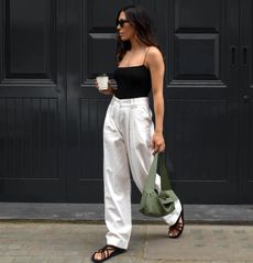 British influencer Jessica Skye poses outside a black paneled building in London wearing black sunglasses, a black tank top with spaghetti straps, white trousers, a green bag, and black strappy flat sandals