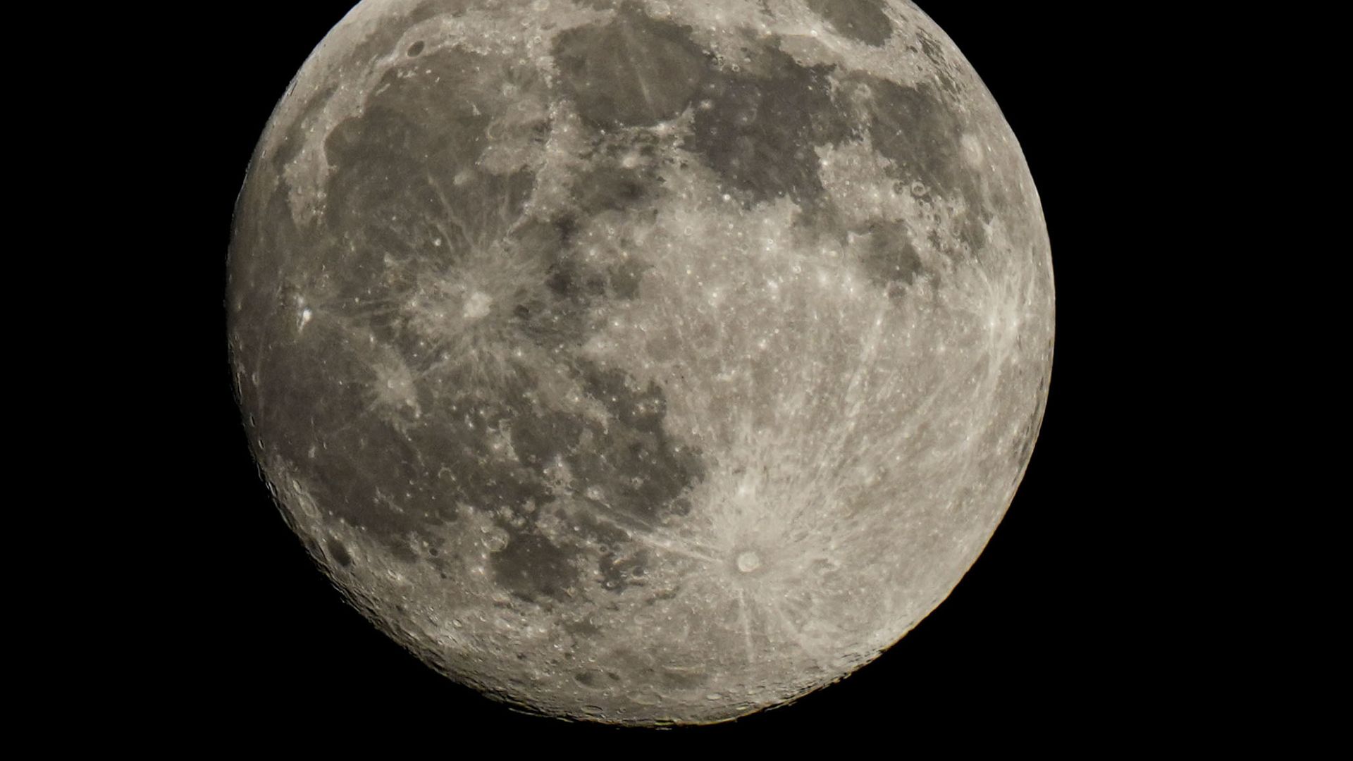 A full moon is photographed in a black night sky with dark lunar seas and bright craters marking its surface.