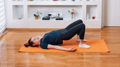 A woman performs a glute bridge on a yoga mat. She is on the ground, with her feet flat against the floor and her knees bent. Her upper body is pushed up off the ground, while her shoulder and arms remain on the floor. Behind her is a white shelf with flowers and photographs.