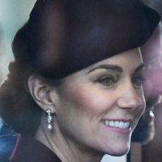 Kate Middleton wearing a maroon hat and coat with diamond earrings riding in a carriage next to Prince William