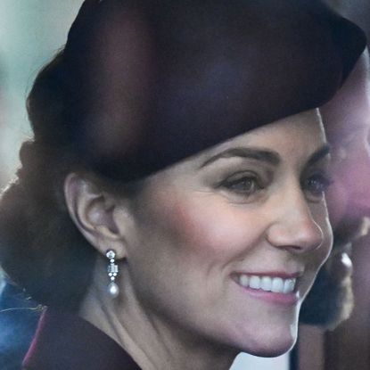 Kate Middleton wearing a maroon hat and coat with diamond earrings riding in a carriage next to Prince William