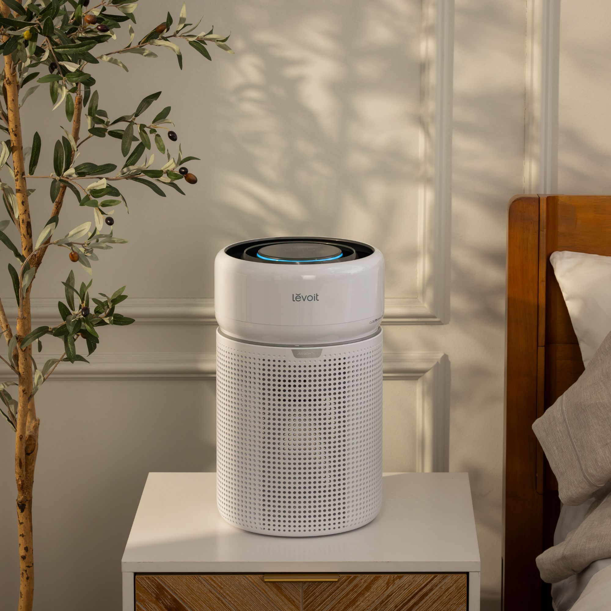 Levoit Eleva 300X Smart Air Purifier sitting on a bedside table, next to a bed and an olive tree