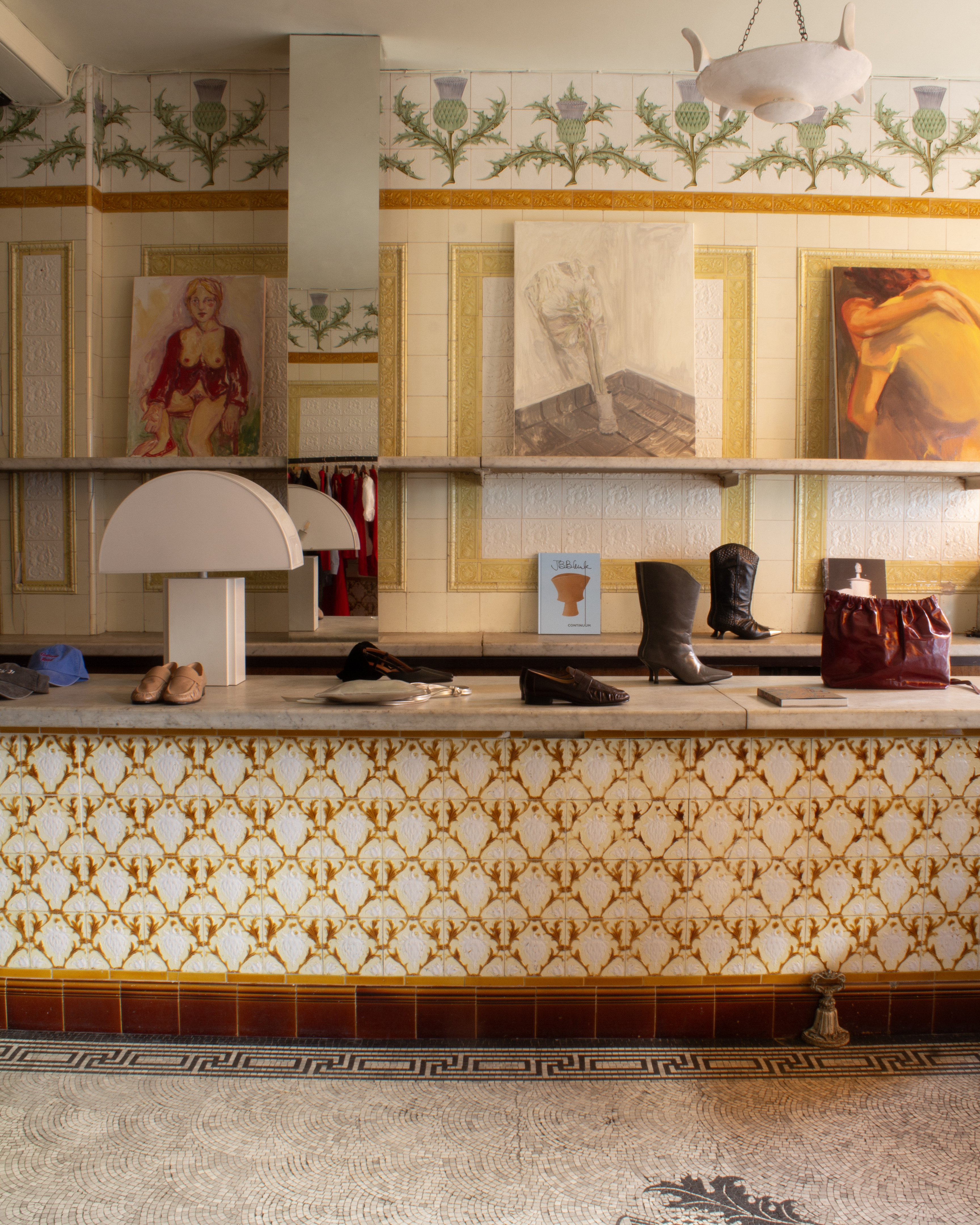 A fashion, design, and art store features historical tiled floors, counters, and walls, ranging from monochrome patterns to terracotta reds, ochres, and vegetal motifs, a marble-topped display of shoes, bags, books, and designer lamps, and a series of figurative canvases.