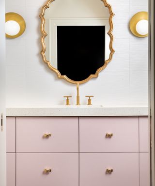 White bathroom with pastel pink cabinetry, brass fixtures and fittings, mirror, pair of wall lights, white counterto