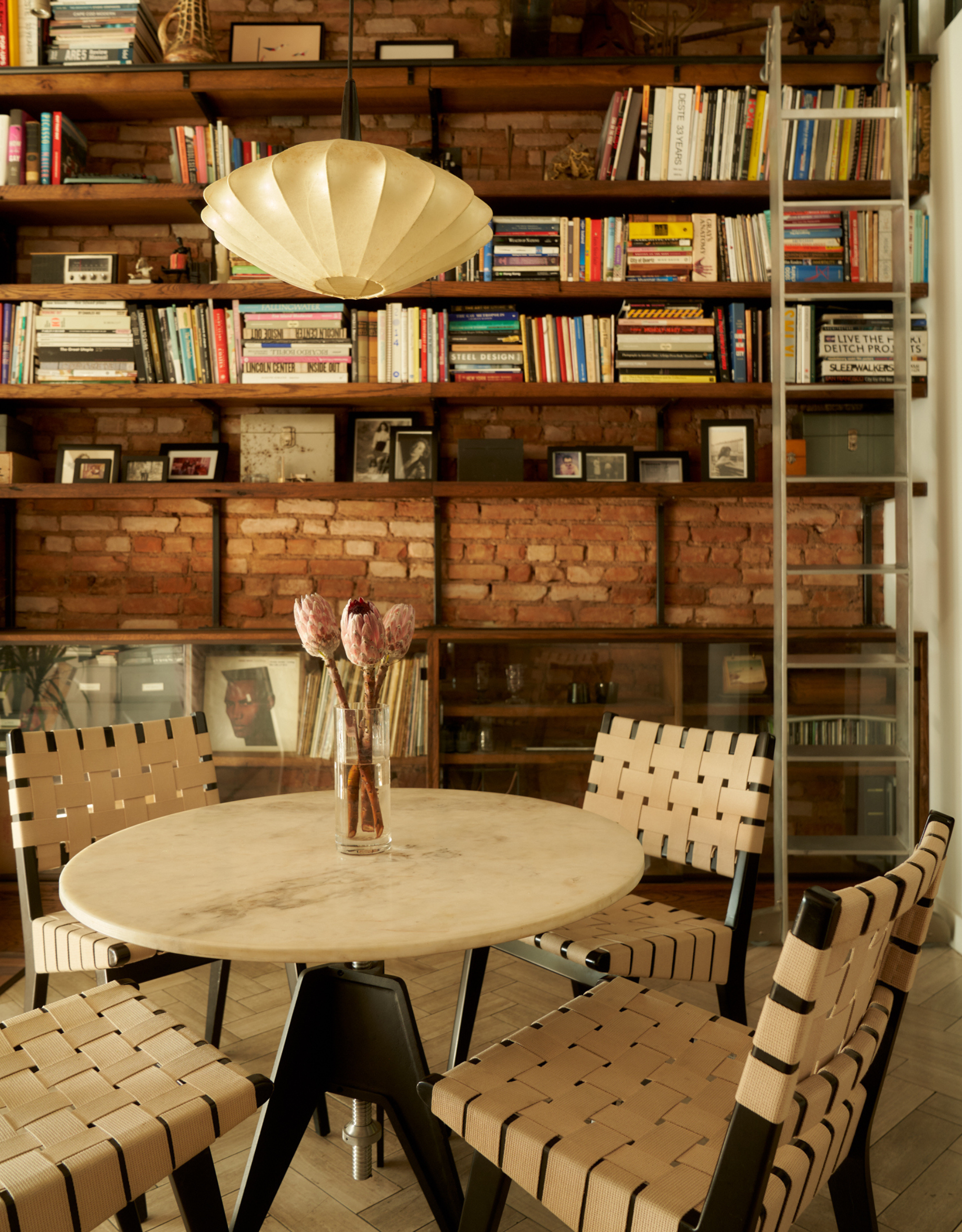 A dining room in an NYC loft with a pendant light, a round stone table with a vase of flowers, a set of black chairs with cream leather detailing, and a bookshelf against a brick wall